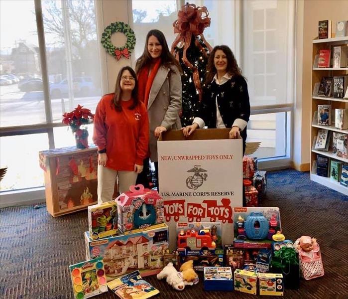 Three individuals in front of Christmas Tree with Toys for Tots box and toys in front of them