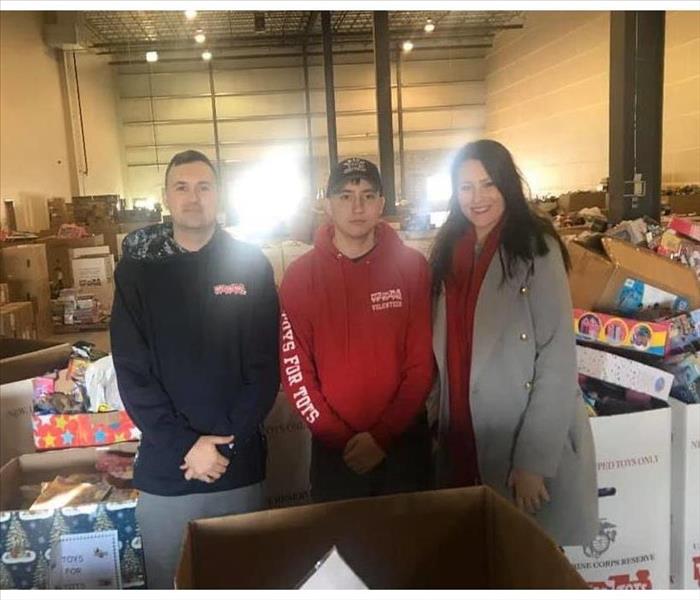 Three individuals in Toys for Tots warehouse with Toys in boxes